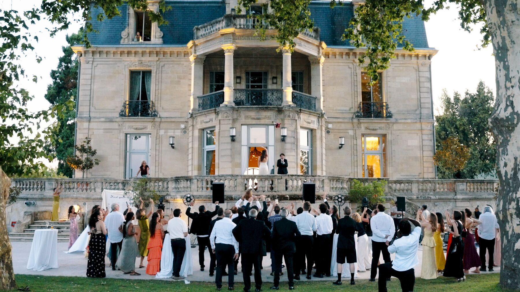 Les mariés au balcon d’un château, lancer de bouquet, invités rassemblés en contrebas.