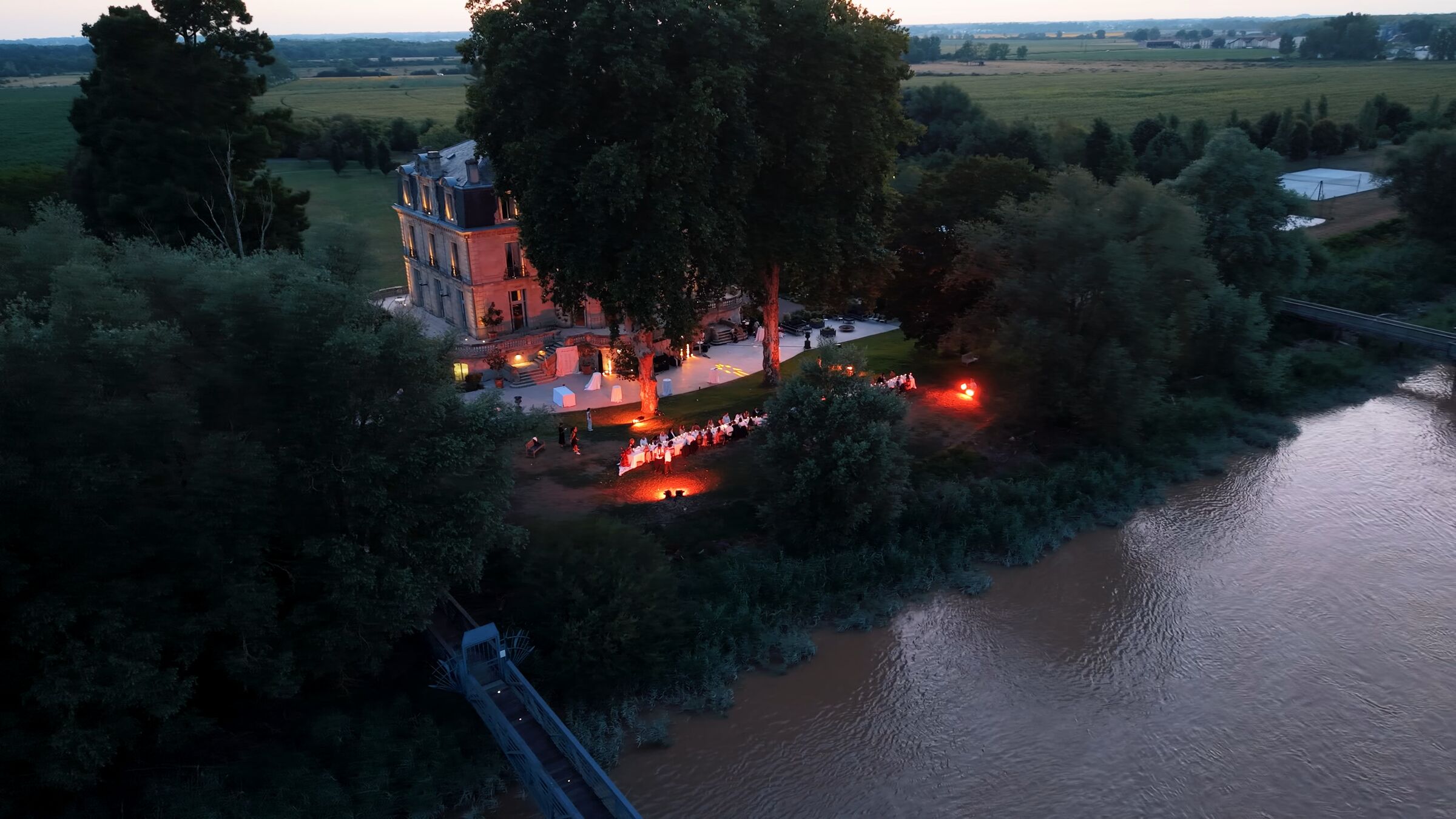 Vue aérienne d’un dîner de mariage au bord de l’eau, dans le parc illuminé d’un château, au crépuscule.